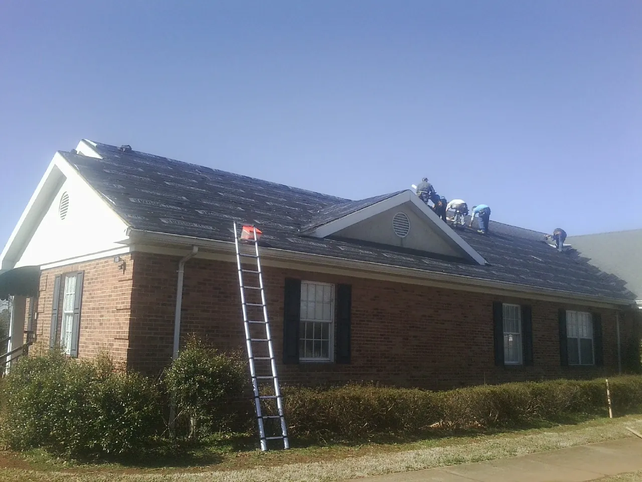 Crew tearing shingles off a brick ranch roof with a ladder set — Metal Roof Repair work by our crew in Raymond